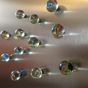 Jeder Murmelmagnet besteht aus einer echten, farbenfrohen Glasmurmel, die an der Rückseite mit einem hochwertigen Neodym-Magneten versehen ist. Die Murmel ist sorgfältig geschliffen, sodass sie eine glatte, angenehm abgerundete Oberfläche hat. Sie fühlt sich kühl und fest an, mit einer sanften Wölbung auf der Vorderseite.

Die Farben der Murmeln variieren – manche sind durchsichtig mit schimmernden Wirbeln, andere sind opalartig, marmoriert oder intensiv gefärbt.