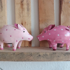 Könnte beinhalten: Zwei dekorative Schweinefiguren. Eines ist hellrosa mit dunkelrosa Punkten, das andere ist dunkler rosa mit weißen Punkten. Beide haben kleine Ohren, Schnauzen und Beine. Sie werden auf einem Holzregal ausgestellt.