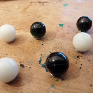 K&ouml;nnte beinhalten: Sechs runde Schrankkn&ouml;pfe. Drei sind gl&auml;nzend wei&szlig; und drei sind schwarz. Jeder Knopf ist auf einer kleinen, silberfarbenen Basis montiert. Die Kn&ouml;pfe sind auf einer hellen Holzoberfl&auml;che angeordnet.