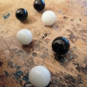 K&ouml;nnte beinhalten: Sechs gl&auml;nzende Murmeln in Schwarz und Wei&szlig; liegen auf einer Holzoberfl&auml;che. Die Murmeln sind kugelf&ouml;rmig und reflektieren das Licht. Die Anordnung deutet auf ein Spiel oder eine dekorative Pr&auml;sentation hin.