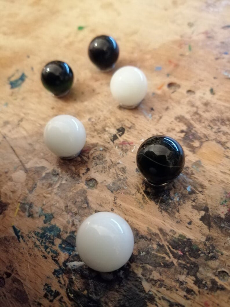 K&ouml;nnte beinhalten: Sechs gl&auml;nzende Murmeln in Schwarz und Wei&szlig; liegen auf einer Holzoberfl&auml;che. Die Murmeln sind kugelf&ouml;rmig und reflektieren das Licht. Die Anordnung deutet auf ein Spiel oder eine dekorative Pr&auml;sentation hin.
