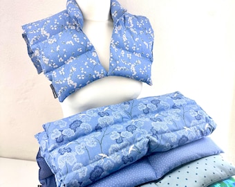 Puede incluir: Un protector de cuello acolchado azul con un patrón de flores de cerezo blancas sobre un maniquí, acompañado de una pila de almohadillas térmicas rectangulares de tela con varios patrones florales, de puntos y geométricos en tonos azules.