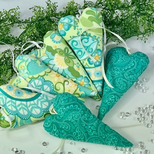 Könnte beinhalten: Ein Set aus sechs herzförmigen Stoff-Anhängern in Grün und Blau mit weißen Schleifen. Die Herzen sind mit floralen und Paisley-Mustern verziert.
