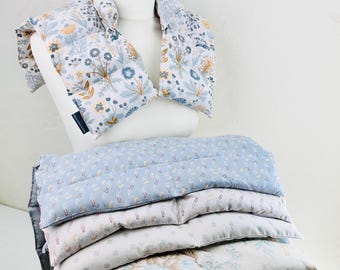 Puede incluir: Un cojín térmico para cuello y hombros de tela blanca con estampado floral en azul, amarillo y gris, sobre un maniquí. Debajo, una pila de cinco almohadillas térmicas rectangulares de tela con varios patrones florales en azul y blanco.