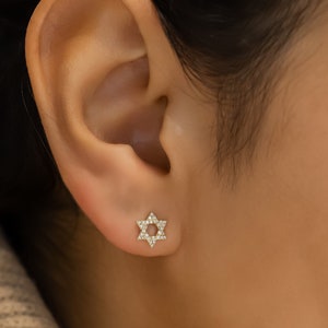 May include: A close-up of a person's ear with a small gold star-shaped earring with diamonds. The earring is a simple and elegant design.