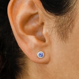 Pode incluir: Um close-up de uma orelha com um pequeno brinco redondo de pedra azul com um halo de pedras brancas menores.
