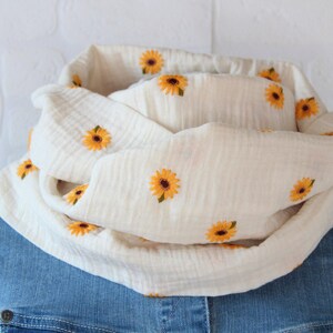 Könnte beinhalten: Ein cremefarbener Rundschal mit gestickten gelben Sonnenblumen. Der Schal ist aus einem weichen, strukturierten Stoff gefertigt und über einer blauen Jeans drapiert. Die Sonnenblumen haben braune Zentren und grüne Blätter.