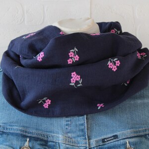 Könnte beinhalten: Ein marineblauer Loop-Schal mit gestickten rosa Blütenzweigen. Der Schal ist über einer Jeansjacke drapiert, wodurch ein mehrschichtiger Look entsteht. Der Schal ist aus weichem, leichtem Stoff gefertigt.