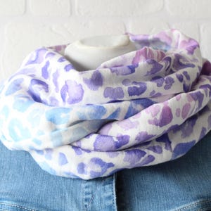 Könnte beinhalten: Ein Loop-Schal mit weißem Grund und einem Leopardenmuster im Aquarell-Stil in Violett-, Lavendel- und Hellblautönen. Der Stoff ist in mehreren weichen Falten drapiert, wodurch eine voluminöse, kreisförmige Form um den Halsbereich entsteht.