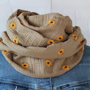 Könnte beinhalten: Ein beigefarbener Loop-Schal mit gestickten gelben Sonnenblumen. Der Schal aus weichem, strukturiertem Stoff ist über einer Jeansjacke drapiert. Die Sonnenblumen haben braune Zentren und grüne Stiele.