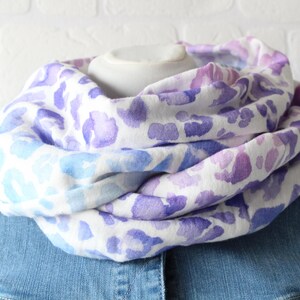 Könnte beinhalten: Ein weißer Loop-Schal mit einem Aquarellmuster aus lila und blauen Flecken. Der Schal ist über einem weißen Schaufensterkopf und einer Jeansjacke drapiert.