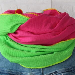 Könnte beinhalten: Ein lebendiger Loop-Schal in leuchtendem Pink und Neongrün. Der Schal ist aus weichem, strukturiertem Stoff gefertigt und mit gelben Nähten versehen. Der Schal ist um eine Jeansjacke gewickelt.