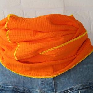 Könnte beinhalten: Leuchtend oranger Schal mit gelben Nähten. Der Schal aus weichem, strukturiertem Stoff ist um den Hals drapiert. Er wird mit einer Jeansjacke getragen.