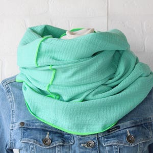 Könnte beinhalten: Ein mintgrüner Loop-Schal mit hellgrünen Nähten ist über einer Jeansjacke drapiert. Der Schal ist aus einem weichen, strukturierten Stoff gefertigt. Die hellgrünen Nähte bilden einen Kontrast zur Farbe des Schals.