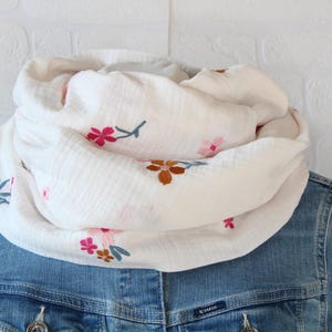 Könnte beinhalten: Weißer Loop-Schal mit gestickten rosa und braunen Blumenmustern. Der Schal besteht aus einem weichen, strukturierten Stoff und wird über einer Jeansjacke getragen. Ein stilvolles Accessoire für jedes Outfit.