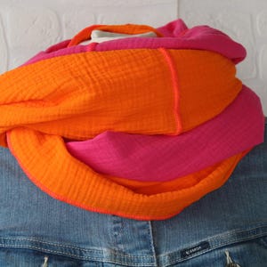 Könnte beinhalten: Ein mehrfarbiger Loop-Schal in Orange und Pink. Der Schal aus strukturiertem Stoff wird über einer Jeansjacke getragen. Die leuchtenden Farben schaffen ein auffälliges Accessoire. Der Schal hat einen Umfang von ca. 150 cm.
