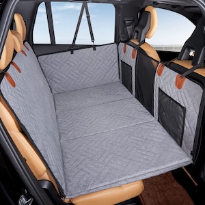 May include: A gray quilted car seat cover designed to protect the back seat of a vehicle. The cover has a diamond pattern and brown leather accents. It is installed in a car with tan leather seats.