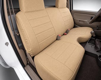 May include: A tan-colored bench seat cover installed in a vehicle interior. The cover features a textured fabric with horizontal stitched lines across the backrests and the seat cushion, designed to fit a split-bench configuration with headrests.