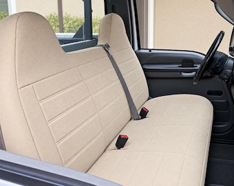May include: A beige fabric bench seat cover installed in a vehicle interior. The cover features a textured weave, horizontal stitched lines, and integrated openings for the seat belt buckles and the shoulder harness strap.