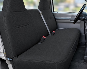 May include: A solid black bench seat cover installed in a vehicle interior. The cover features a textured fabric surface with integrated headrests, visible seat belt buckles, and a structured design that contours to the shape of the bench seat.