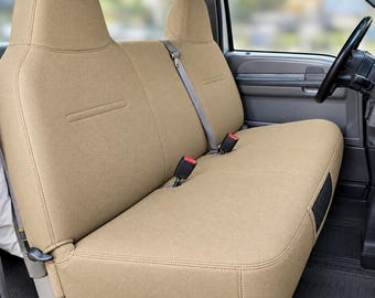 May include: A tan-colored bench seat cover installed in a vehicle interior. The cover features a textured fabric, integrated headrests, and openings for seat belts with red release buttons. The design includes visible stitching along the seams.