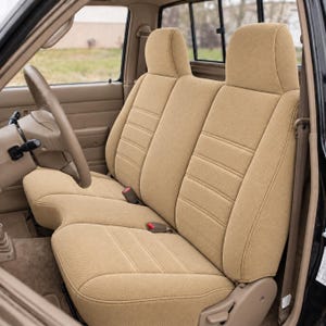 May include: A tan fabric seat cover set installed on a vehicle bench seat. The covers feature a textured weave pattern with horizontal stitched lines across the backrests and seat cushions, including integrated headrests and seat belt buckles.