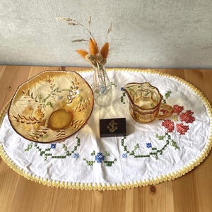 Könnte beinhalten: Ein ovales weißes Stoffdeckchen mit gelbem Häkelrand und floraler Kreuzstichstickerei, darauf eine bernsteinfarbene Glasschale, ein kleiner bernsteinfarbener Glaskrug, eine klare Glasvase mit Trockenblumen und eine kleine dunkle Holzschatulle mit Anker.