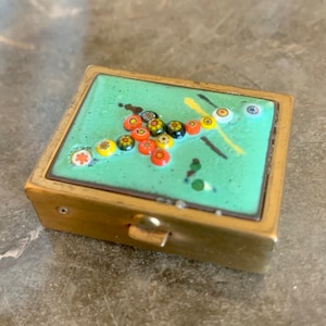 May include: A small, square, brass trinket box with a hinged lid. The lid is decorated with a green enamel background and a colourful mosaic of small, round glass pieces in shades of red, yellow, orange, black, and white.