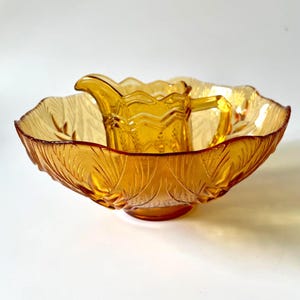 Könnte beinhalten: Bernsteinfarbenes Glasschalen- und Krug-Set. Die Schale hat ein Blattmuster am Rand und der Krug hat einen Griff. Das Set besteht aus Glas und ist wahrscheinlich Vintage. Der Krug sitzt in der Schüssel.