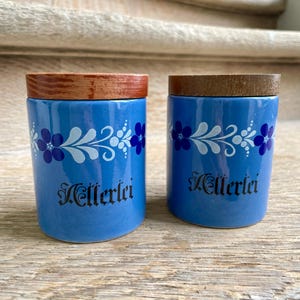 May include: Two matching blue ceramic jars with wooden lids. Each jar features a floral design in white and blue, with the word "Kellerlei" in black script. The jars are cylindrical and sit on a wooden surface.