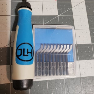 Puede incluir: Un cuchillo artesanal con mango azul y blanco y un juego de cuchillas de repuesto en una caja de plástico transparente. El mango tiene las letras "JLH" dentro de un círculo azul.