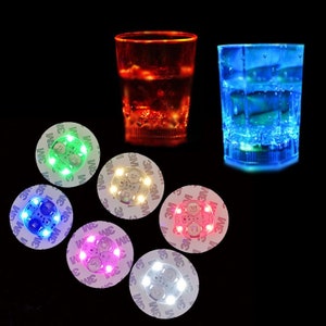 Könnte beinhalten: Eine Sammlung von LED-Leuchtuntersetzern und zwei Schnapsgläsern. Die Schnapsgläser leuchten, eines rot und eines blau. Die Untersetzer haben verschiedenfarbige Lichter, darunter grün, blau, weiß, rosa und rot.