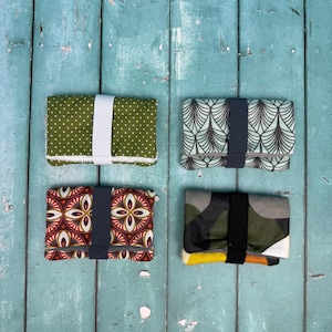 Może przedstawiać: Cztery materiałowe portfele z gumkami. Jeden jest oliwkowozielony z białymi kropkami i białą gumką. Inny ma turkusowo-czarny wzór art deco z granatową gumką. Trzeci ma czerwony, brązowy i złoty wzór kwiatowy z granatową gumką. Ostatni ma wzór kamuflażu z czarną gumką.