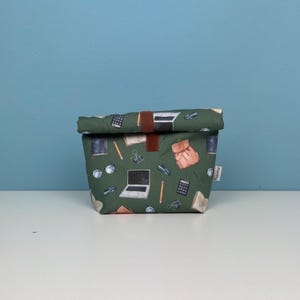 Könnte beinhalten: Eine grüne Lunch-Tasche mit einem Aufdruck von Schulutensilien, darunter ein Laptop, Bleistifte, Brillen und ein Rucksack. Die Tasche hat einen braunen Lederriemen.
