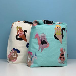 May include: Two fabric tote bags with a whimsical design. One bag is off-white, the other is light blue. Both bags feature illustrations of women in swimsuits on inflatable swans and unicorns. The bags are on a white surface with a blue background.