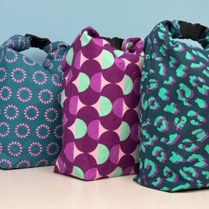 Könnte beinhalten: Drei farbenfrohe, gemusterte Taschen mit schwarzen Griffen. Die Taschen zeigen Türkis mit rosa Blumendesigns, Lila mit rosa und mintfarbenen geometrischen Formen und Türkis mit grünem und lila Leopardenmuster. Die Taschen stehen aufrecht.