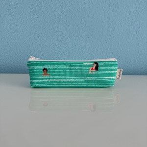 Może przedstawiać: Turkusowy, zapinany na zamek błyskawiczny, piórnik z białym tłem i wzorem białych pasków. Piórnik przedstawia dwie postaci ludzi pływających w wodzie. Na boku piórnika znajduje się napis "Watertour".