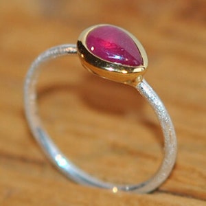 Puede incluir: Anillo de plata con una gema roja oscura en forma de lágrima engastada en un bisel dorado. La banda del anillo tiene un acabado texturizado. La gema es el punto focal de la joya.
