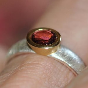 Op de afbeelding: Een ring met een ronde, dieprode edelsteen in een gouden zetting. De band is gemaakt van getextureerd zilver met een matte afwerking, wat zorgt voor een contrast tussen de gladde gouden zetting en het ruwe metalen oppervlak van de ring.