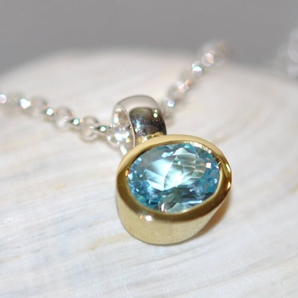 May include: A pendant necklace featuring an oval-cut light blue gemstone set in a smooth yellow gold bezel. The pendant is attached to a delicate silver chain, which connects to the top of the gold setting with a polished silver bail.