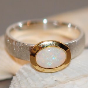 Può includere: Un anello con una fascia in argento testurizzato e una pietra di opale bianco di forma ovale al centro. L'opale è incastonato in una lunetta in oro liscio e lucido, creando un contrasto tra la finitura opaca dell'argento e l'oro.