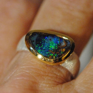 May include: A silver ring with a gold bezel holding an iridescent opal gemstone. The opal shows blues and greens. The ring is on a finger. The ring's band is silver.