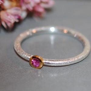 May include: A silver ring with a textured band and a small, round, pink gemstone set in a gold bezel. The ring is displayed on a gray surface, with pink flowers in the background.