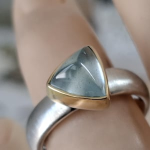 Op de afbeelding: Zilveren ring met een driehoekige, lichtblauwe edelsteen gezet in een gouden bezel. De ringband heeft een geborstelde metalen afwerking, de edelsteen is doorschijnend.