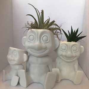 以下が含まれることがあります： 漫画のキャラクターの形をした白いセラミック製のプランターが3つあり、それぞれに異なる植物が植えられています。一番大きなプランターには緑色のエアプラント、真ん中のプランターには緑色の多肉植物、一番小さなプランターは空です。