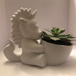 Könnte beinhalten: Weißer Keramik-Einhorn-Pflanztopf mit einer grünen Sukkulentenpflanze im Inneren. Das Einhorn hält den Topf mit seinen Vorderbeinen.