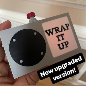 Può includere: Un dispositivo rettangolare grigio con un componente circolare nero e un pomello rosso. Il testo "WRAP IT UP" è visualizzato su uno sfondo rosa. È visibile anche la frase "New upgraded version!"