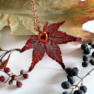 Puede incluir: Un collar de color cobre con un colgante de hoja de arce roja y plateada detallada. El colgante cuelga de una cadena y está rodeado de una hoja marrón y bayas negras.