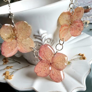 May include: A delicate necklace featuring three pink flower charms with a silver chain. The flowers have a translucent, almost iridescent quality, and are accented with glitter.