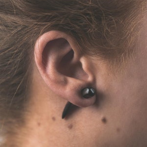 May include: Close-up of an ear with a dark, teardrop-shaped earring. The earring is a dark, solid color, possibly black or a very dark brown. The skin around the ear is light-toned, with a few freckles visible.